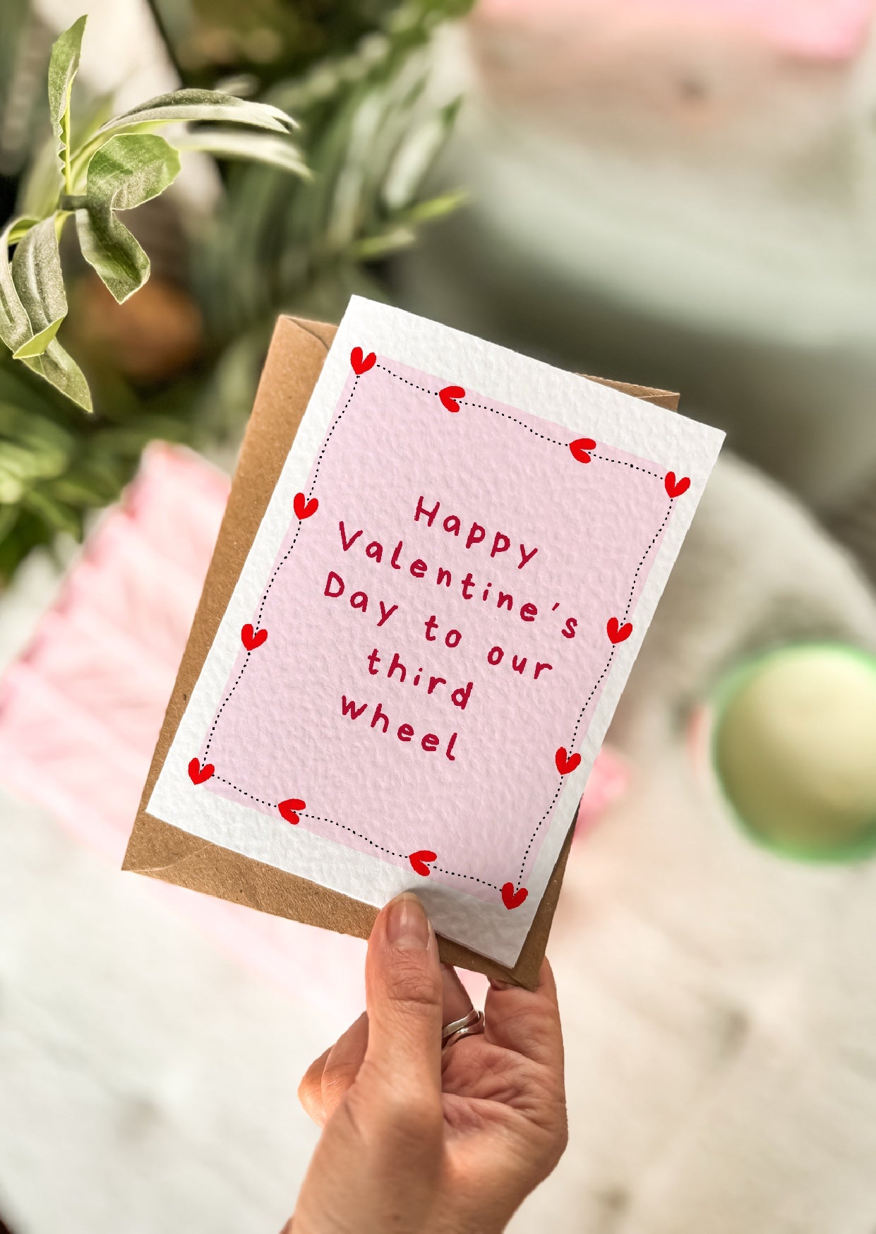 Hand holding a Valentine's Day card with 'Happy Valentine's Day to our third wheel' text.